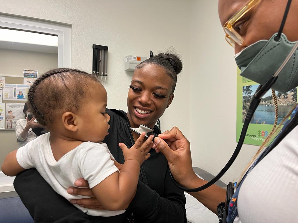 A Black doctor with blonde and black box braids using a stethoscope on a Black baby. The baby is held by a smiling Black mom.